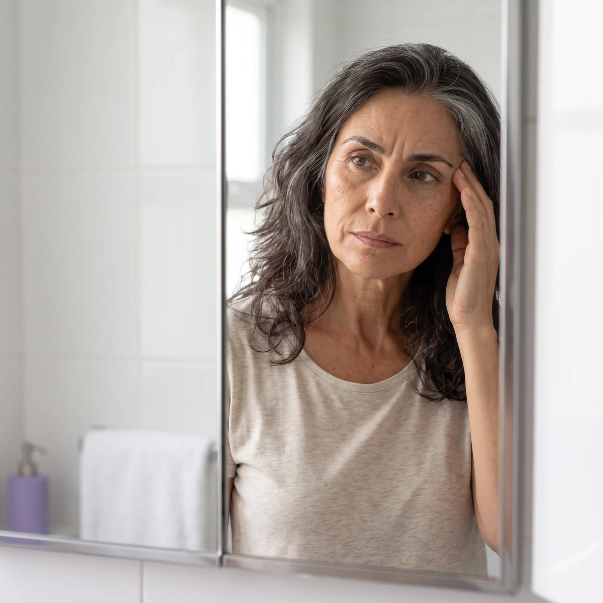 Mujer de unos 50 años frente al espejo del baño observando la línea de expresión de su entrecejo